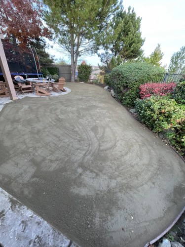 Backyard patio with a circular seating area around a stone fire pit, trampoline in view, and trees and hedge edging the yard.