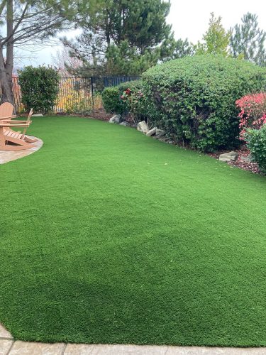 Artificial turf lawn with two peach-colored Adirondack chairs on a concrete pad, bordered by shrubs and a black metal fence.